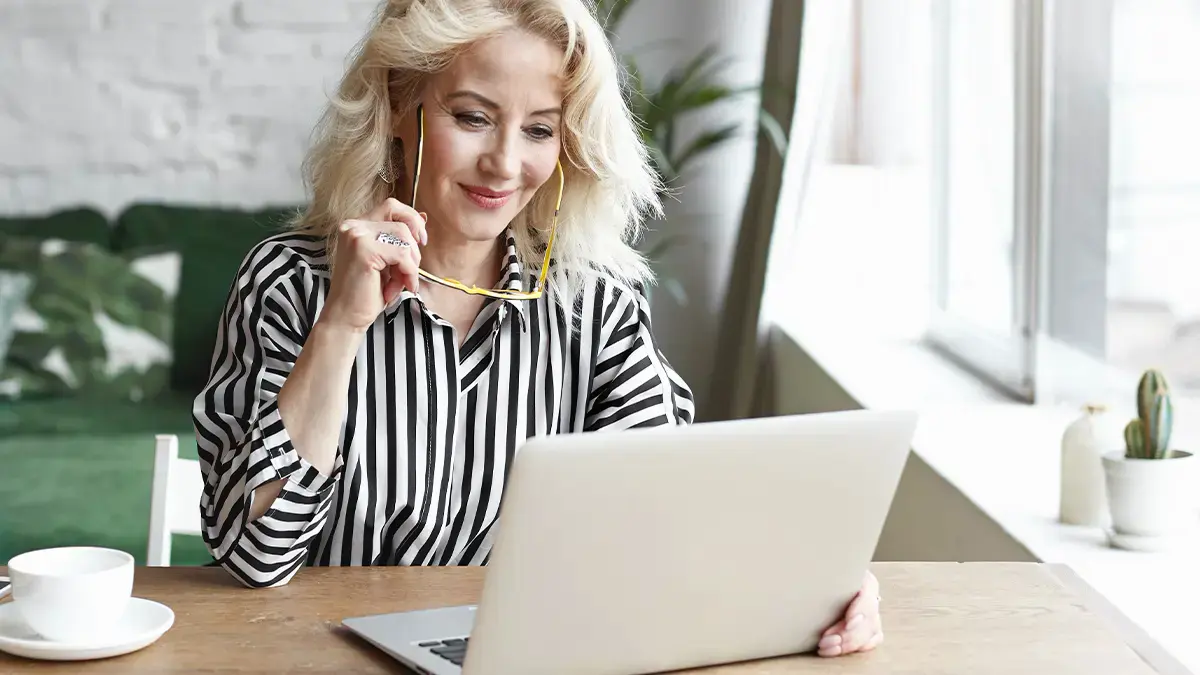 Woman holding her glasses and smiling at laptop in home office